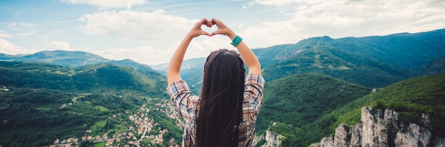 girl standing on a hilltop overlooking lots if small houses, making a heart shape with her hands