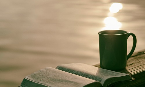3-landing a coffee mug and a bible sit on a wooden table in front of a lake with the reflection of the sunrise