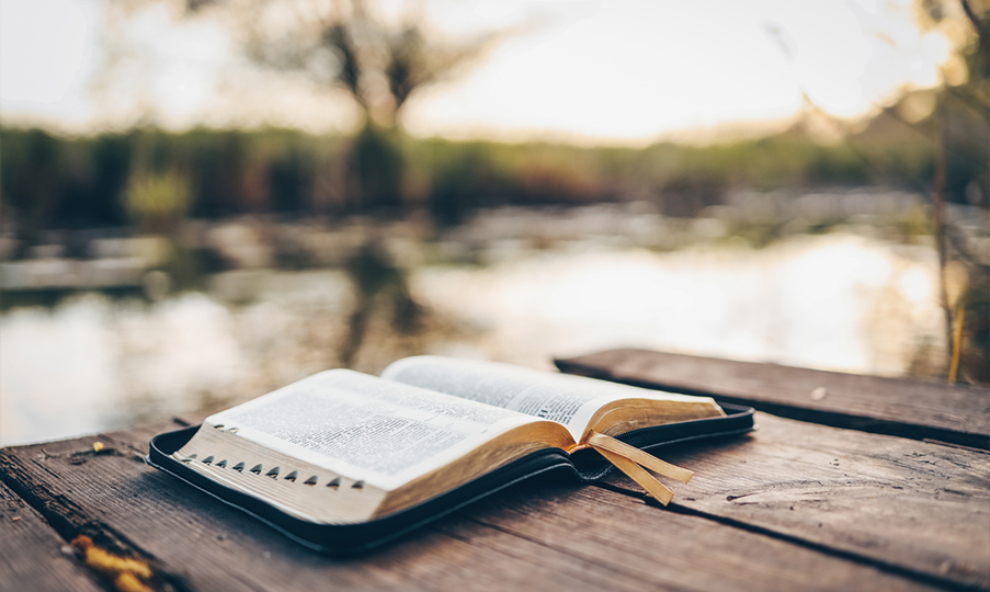 an open bible sitting on a wooden table in front of a calming and still lake