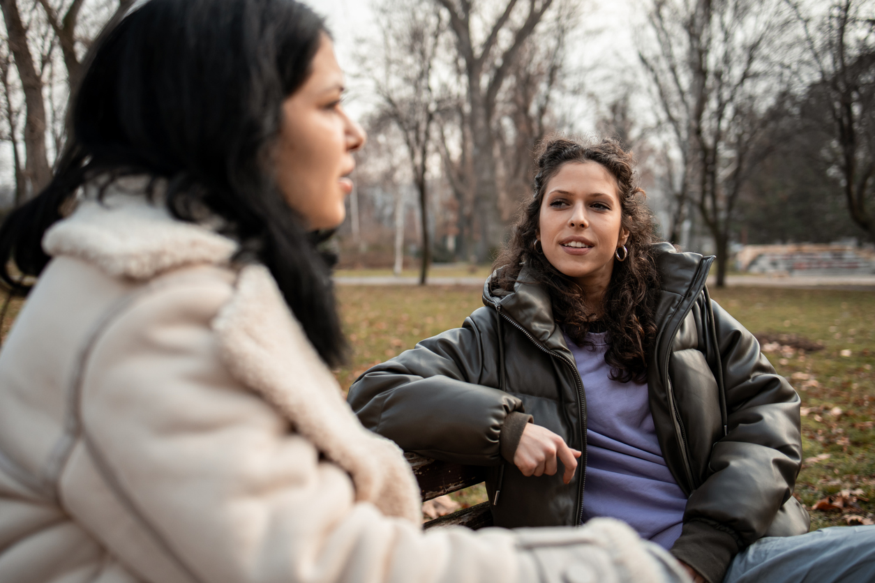 Two women talking on a park bench in winter, with one woman in focus listening while the other speaks in the foreground.