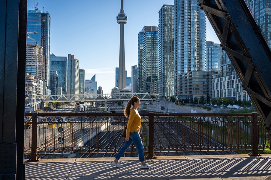 2 - For the Web - Editorial - iStock-1469320186 A woman in a yellow sweater walks across a overpass bridge, Toronto's CN tower is in the background